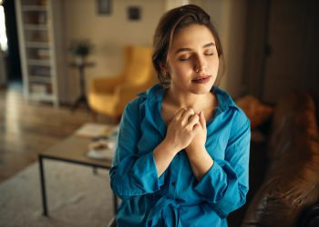 Portrait of joyful charming young female sitting on sofa in living room closing eyes, keeping hands on her chest, praying, having calm peaceful facial expression, dreaming of something pleasant
