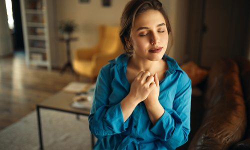 Portrait of joyful charming young female sitting on sofa in living room closing eyes, keeping hands on her chest, praying, having calm peaceful facial expression, dreaming of something pleasant