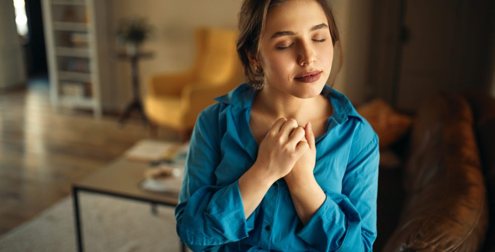 Portrait of joyful charming young female sitting on sofa in living room closing eyes, keeping hands on her chest, praying, having calm peaceful facial expression, dreaming of something pleasant